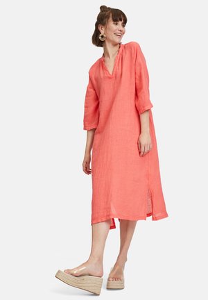 Korallenfarbenes Leinenkleid mit Dreiviertelärmeln, V-Ausschnitt, seitlichen Schlitzen und lockerer Passform. Model kombiniert es mit transparenten, hohen Sandalen.