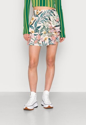 Kvinde iført en lagdelt miniskørt med blomsterprint, grønstribet langærmet crop top og hvide høje sneakers, stående på en grå gulv.