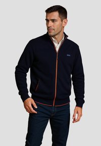 Navy zip-up sweater met oranje accenten, voorzien van een hoge kraag, geribbelde manchetten en zoom, en twee zijzakken. Gemaakt van een zachte, gestructureerde stof.