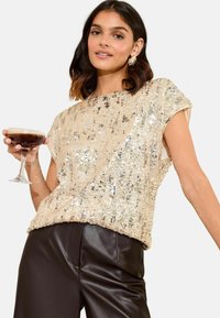 Beige sequin-pitsi yllärillä, joissa on lyhyt hiha ja löysä malli. Yhdistetty ruskeisiin nahkahousuihin ja pidetään cocktaillasia.