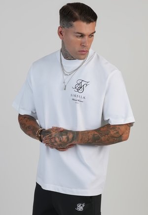 Camiseta blanca de manga corta con cuello redondo, que presenta un logo de marca y texto en negro. Fabricada en suave algodón, con un corte relajado, combinada con pantalones negros.