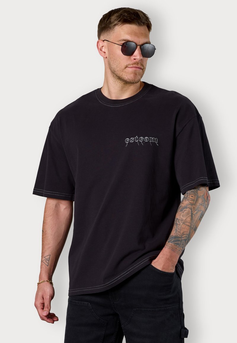 Black oversized t-shirt made of cotton with contrast stitching. Features a small embroidered logo on the chest. Worn with sunglasses.
