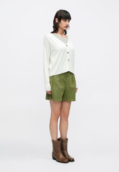 Γυναίκα που φοράει λευκό ζακέτα με κουμπιά, γκρι μπλουζάκι, πράσινες σορτς και καφέ μπότες μέχρι τη μέση της γάμπας, στέκεται μπροστά σε απλό λευκό φόντο.