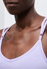 Κοντινό πλάνο ενός ατόμου που φοράει ένα ανοιχτό μωβ τοπ με ρυθμιζόμενους ιμάντες και με μια αχνή τατουάζ ορατή στο επάνω μέρος του στήθους.