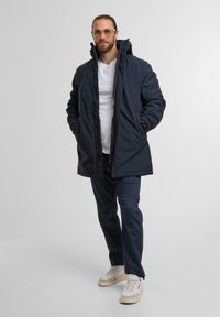 Giacca isolante blu navy con cappuccio, dotata di chiusure a zip e bottoni, indossata su una maglietta bianca, abbinata a pantaloni scuri e scarpe da ginnastica chiare.