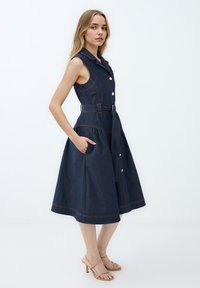 Dunkelblaues, ärmelloses Kleid aus Denim mit einem Gürtel in der Taille, großen Knöpfen, seitlichen Taschen und einem ausgestellten Rock. Verfügt über kontrastierende Stickdetails.