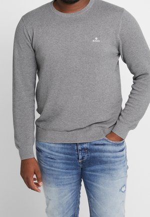 Pull en maille grise avec un col rond et des poignets côtelés, arborant un petit logo blanc sur la poitrine. Associé à un jean en denim bleu délavé.