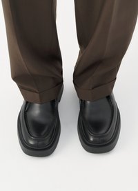 Svarta läderloafers med en kraftig sula, detaljerad sömnad och rund tå, parat med bruna, plisserade byxor.