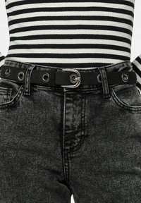 Cinturón de cuero negro con herrajes plateados y ojetes, llevado sobre unos vaqueros de mezclilla gris oscuro, combinado con una blusa a rayas en blanco y negro.
