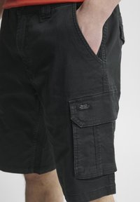 Svarte cargo-shorts laget av stoff med en glatt tekstur. Har sidelommer, en lommepose på låret, og en merking detalj på siden.