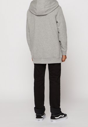 Person iført en grå hættetrøje, sorte bukser og sorte sneakers med hvide såler, stående med ryggen vendt mod en ensfarvet baggrund.