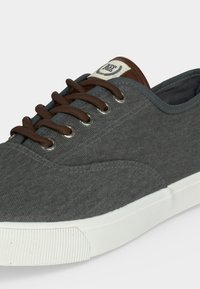 Σκούρα γκρι πάνινο sneaker με καφέ κορδόνια, λευκή ελαστική σόλα και ένα μικρό λογότυπο στη γλώσσα.