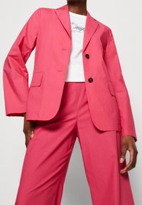 Blazer rose à deux boutons, col cranté, et poches avant, associé à un pantalon large assorti. Texture lisse. Chemise blanche en dessous.