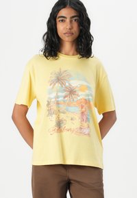 Gul bomull t-shirt med grafisk design av palmer, berg och en figur på en strand. Löst skuren, kortärmad och med rund hals.