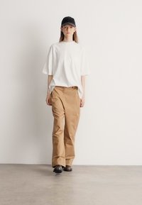 Biała oversize koszulka, beżowe spodnie o szerokich nogawkach i czapka z daszkiem w kolorze czarnym. Model stoi na jednolitym tle, prezentując casualowy, prosty styl.