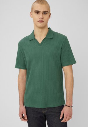 Jeune homme aux cheveux blonds courts portant un polo vert texturé à manches courtes et un jean foncé sur un fond gris uni.