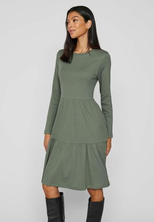Femme aux longs cheveux foncés portant une robe longue jusqu'aux genoux, à manches longues, de couleur vert olive et des bottes noires montantes jusqu'aux genoux, debout devant un fond uni.