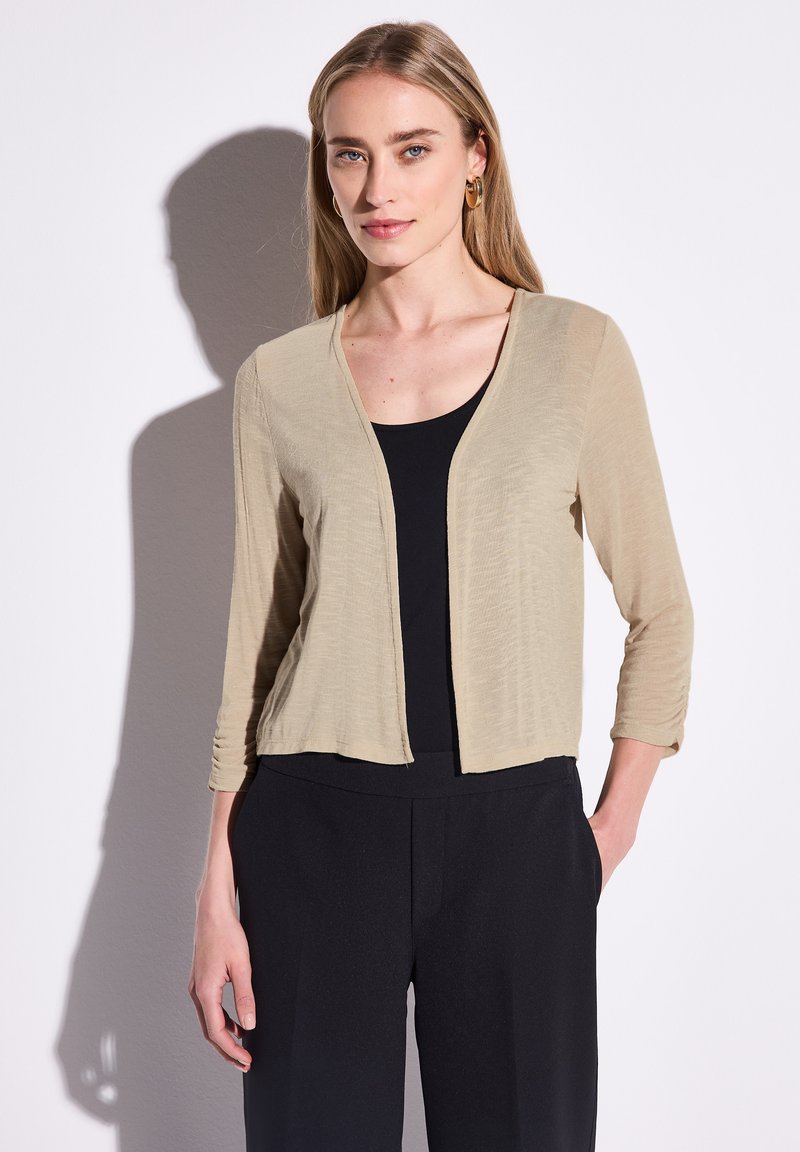 Ljusbeige kort kavaj i texturerad tyg, med V-ringning och trekvartslånga ärmar, bärs över en svart topp.