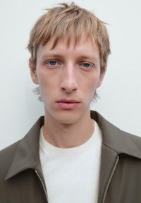 Mand iført en brun zip-up jakke over en hvid crewneck trøje. Han har lysebrunt hår og blå øjne, med et neutralt udtryk.