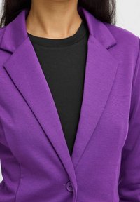 Kvinna i en lila kavaj över en svart rundhalsad tröja, som visar övre delen av överkroppen och halslinjen.