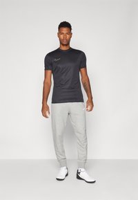 Maglietta atletica nera a maniche corte con logo Nike dorato, abbinata a pantaloni della tuta grigi chiari e sneakers bianche con dettagli neri.