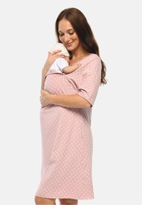 Woman in pink heart-patterned dress gently holding sleeping baby wrapped close to her chest against a plain background.