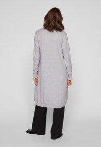 Długi szary kardigan z miękkiego dzianinowego materiału, z prążkowanymi mankietami i dołem, noszony na czarnych spodniach, o prostym, minimalistycznym kroju.