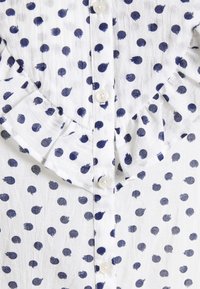 Camisa de tecido branco com bolinhas náuticas, apresentando um colarinho franzido e botões à frente. A textura parece ligeiramente amarrotada.
