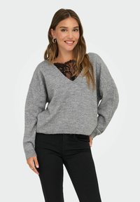 Femme souriante portant un pull gris en V sur un top en dentelle noire, un pantalon noir et des boucles d'oreilles en or, posant avec une main sur la hanche.