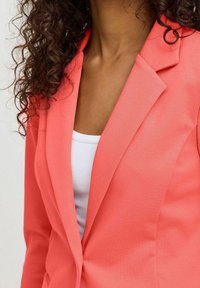 Blazer corail avec un revers cranté, coupe ajustée et texture lisse, porté sur un débardeur blanc.