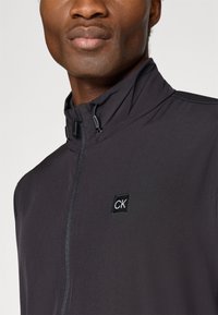 Giacca nera leggera con collo alto, dotata di zip frontale e di un piccolo logo CK sul lato sinistro. Texture liscia e design moderno.