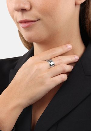 Silberner Ring mit einer polierten Oberfläche und flacher Form, versehen mit einem subtilen eingravierten Detail. Die Hand des Models liegt gegen ihren Kragen.