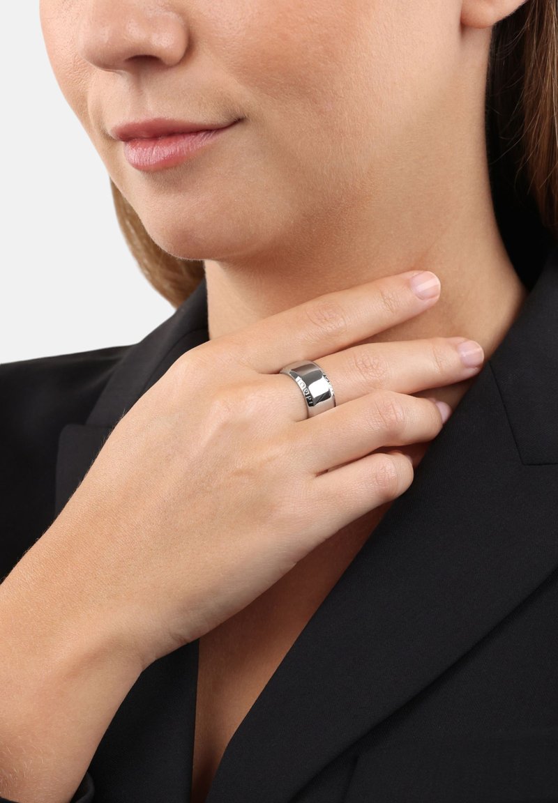 Anello d'argento con una finitura lucida e una superficie piatta, caratterizzato da un delicato dettaglio inciso. La mano della modella è posizionata contro il suo colletto.