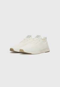White knit sneakers with textured upper, featuring a rounded toe, tonal laces, and a beige rubber sole. Silver accent on the heel.
