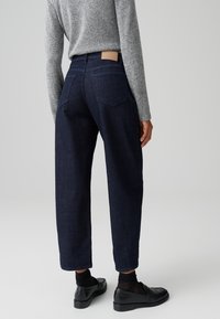 Jeans in denim scuro a vita alta con una vestibilità rilassata, caratterizzati da un'etichetta in pelle beige, due tasche posteriori e orli cuciti. Indossati con un top in maglia grigia.