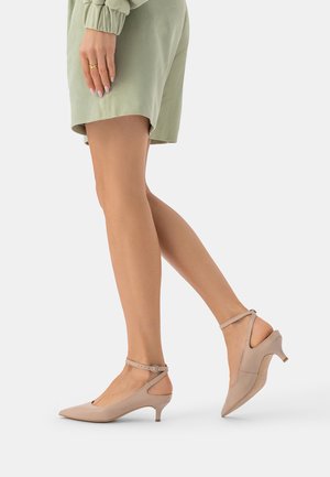 Kobieta nosząca beżowe buty na niskim obcasie, z zaostrzonym noskiem i paskami wokół kostki, w połączeniu z zielonymi szortami szałwiowymi oraz dopasowaną, długorękawową bluzką.