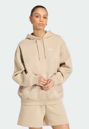 Kobieta w beżowej, oversize bluzie z kapturem Adidas z logo i dopasowanych szortach, stojąca na jednolitym jasnym tle.