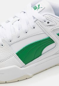 Valkoiset tennarit, joissa on vihreä sivuviiva, valkoiset nauhat ja Puma-logo kielekkeellä, näyttäen ommellun nahkan ja tekstuuripohjan yksityiskohtia.