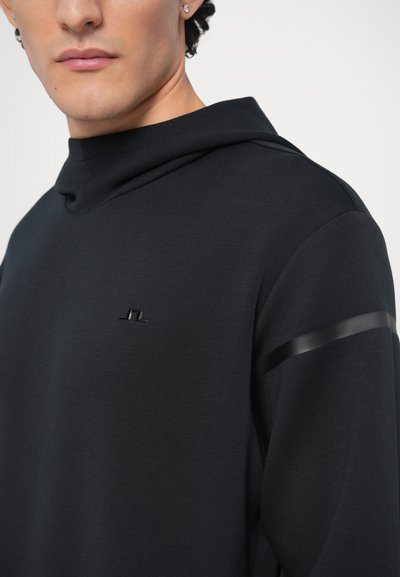 Μαύρη φούτερ με υψηλό γιακά, υφή υφάσματος και διακριτικό λογότυπο στο στήθος. Διαθέτει γυαλιστερή μαύρη ρίγα στο μανίκι.