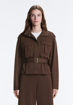 Kobieta o długich włosach, ubrana w brązową kurtkę z paskiem, kieszeniami na klapki z przodu oraz pasujące brązowe szerokie spodnie, stojąca na białym tle.