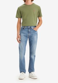 Grön t-shirt i bomull med liten röd logga, kombinerad med ljusblå denimjeans med rak passform. Vita sneakers kompletterar outfiten.