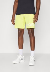 Shorts jaune fluo avec un imprimé blanc "BOR", associés à des chaussures de sport blanches et des chaussettes blanches arrivant à la cheville, portés avec un t-shirt noir.