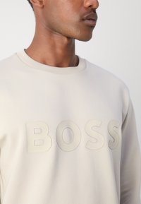 Sudadera de color beige claro con cuello redondo y gran logotipo en relieve "BOSS" en un tono ligeramente más oscuro. Textura de tela suave.