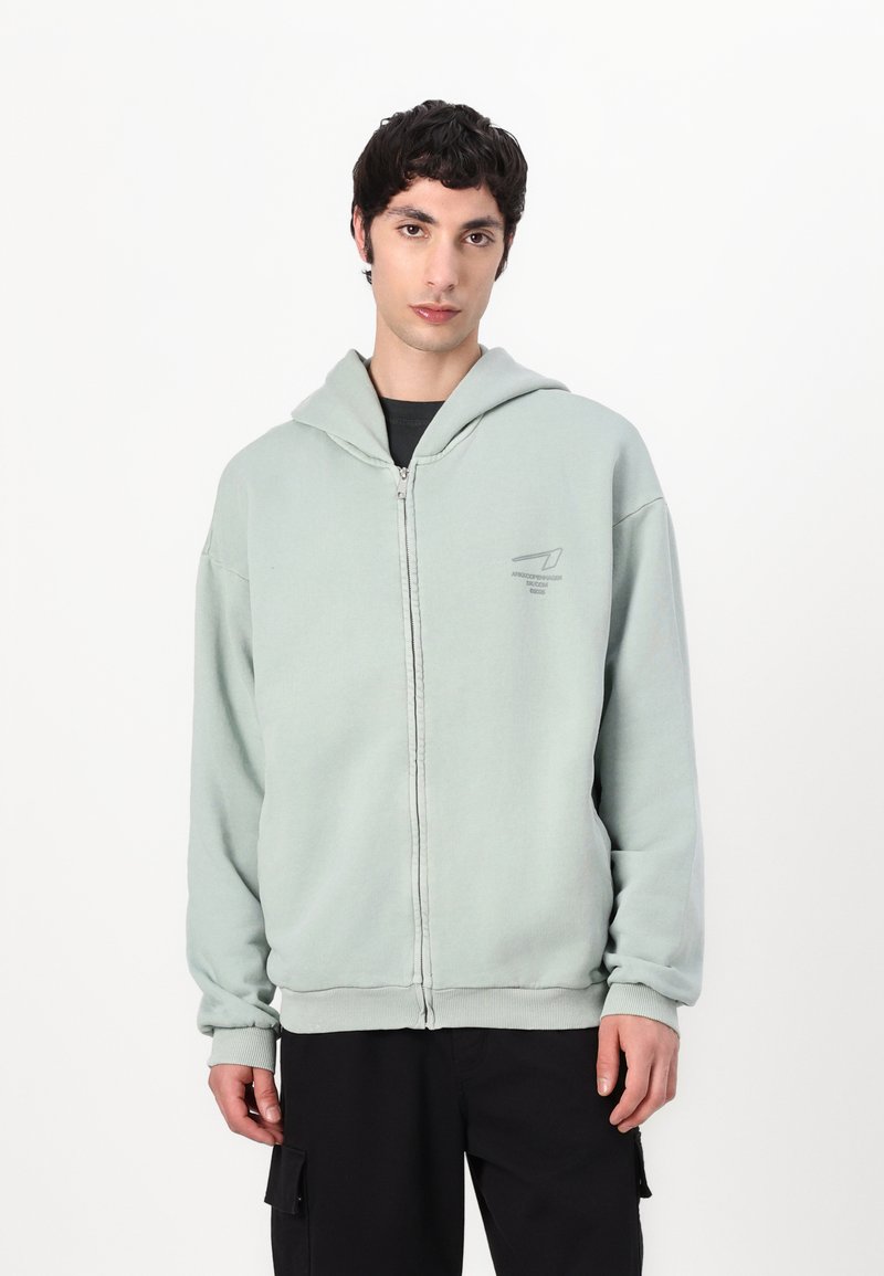 Light green zip-up hoodie with large hood, ribbed cuffs, and hem. Features a subtle graphic on the chest. Worn over a black shirt.