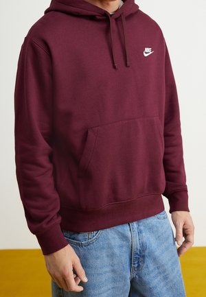 Bordowa bluza z kapturem marki Nike z przednią kieszenią, noszona z jasnoniebieskimi dżinsami, pokazująca tors i górną część nóg.