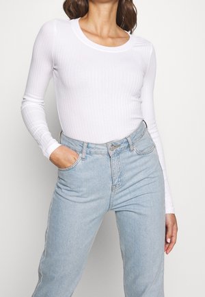 Femme portant un haut blanc côtelé à manches longues rentré dans un jean taille haute bleu clair, posant avec une main dans la poche.