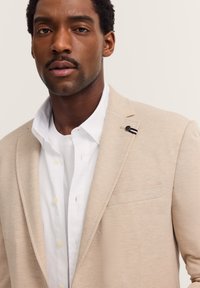 Beige Blazer aus strukturiertem Stoff, mit einer einzigen Brusttasche und schwarzen sowie weißen Akzentdetails. Getragen über einem weißen Hemd mit Kragen.