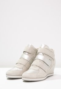 Beżowe sneakersy na koturnie zamszową cholewką i metalicznymi srebrnymi paskami. Z boku zamek błyskawiczny i teksturowany wzór. Gumowa podeszwa zapewniająca przyczepność.