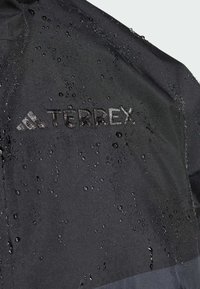 Giacca impermeabile nera con finitura opaca. Presenta un marchio "TERREX" in evidenza, gocce d'acqua sulla superficie e una texture liscia.