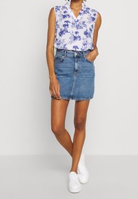 Nainen, jolla on yllään valkoinen, sinisin kukkakuosin varustettu hihaton pusero, sininen farkku-minihame ja valkoiset korolliset tennarit, seisoo käsi taskussa.